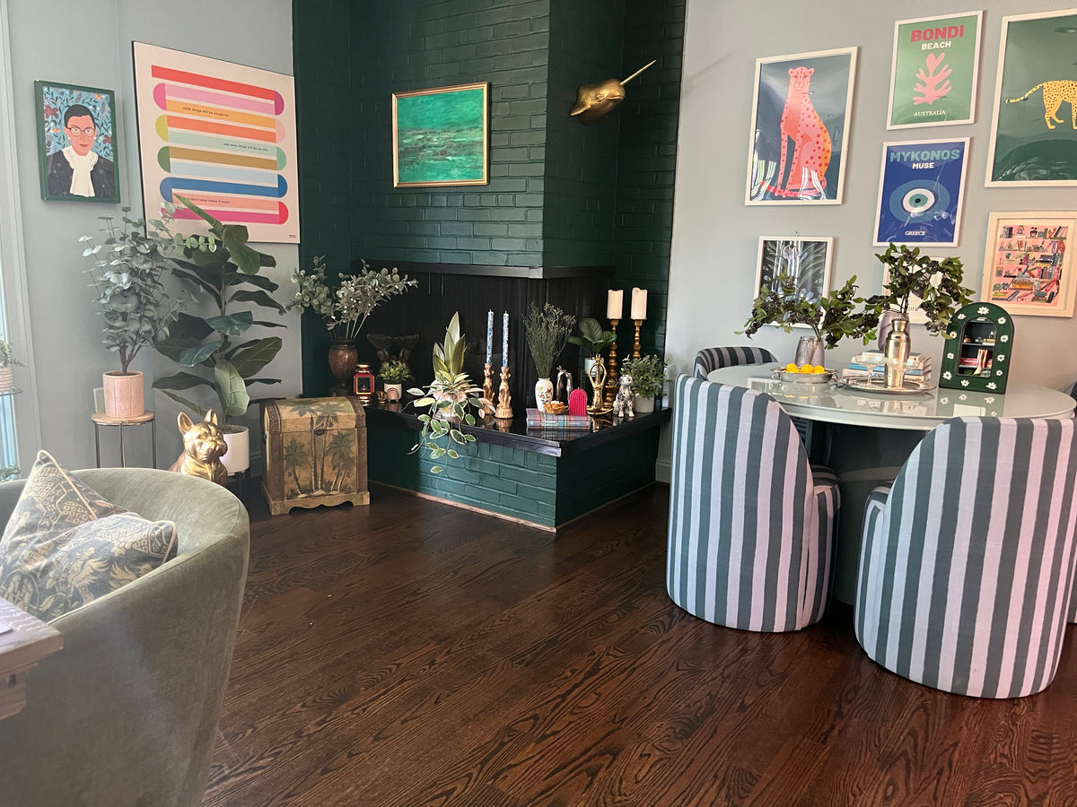 Vintage decor around a dark green brick fireplace, with framed landscape art, a trunk, and striped dining chairs.